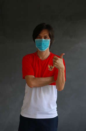 Man wearing hygienic mask and wearing Indonesian flag colored shirt and cross one s arm with thumbs up on dark wall background. Concept of protect tiny dust or disease of Indonesia.の写真素材