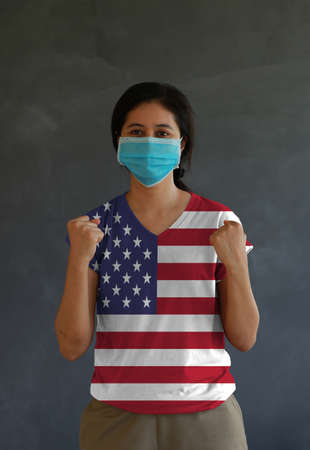 Masked woman wearing USA flag color of shirt and standing with raised both fist on dark wall background. Concept of protection and fighting COVID.の写真素材