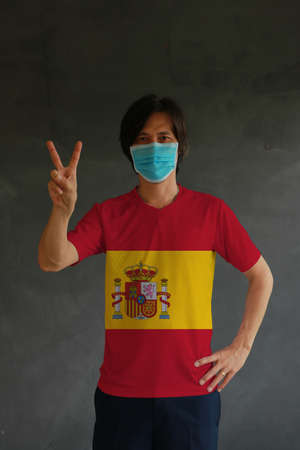 Masked man wearing Spain flag color of shirt and raising two fingers up on dark wall background. Concept of protection and fighting COVID.の写真素材