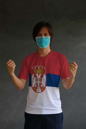 Man wearing hygienic mask and wearing Serbia flag colored shirt and standing with raised both fist on dark wall background. Concept of protect tiny dust or disease.の写真素材