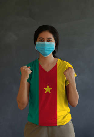 Woman wearing hygienic mask and wearing Cameroon flag colored shirt and standing with raised both fist on dark wall background. Concept of protect tiny dust or disease.の写真素材