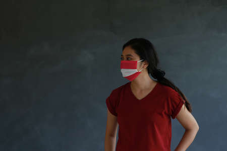 Woman wearing Austrian flag colored hygienic mask with brown shirt and facing to the side on dark wall background. Concept of protect tiny dust or disease of Austria.の写真素材