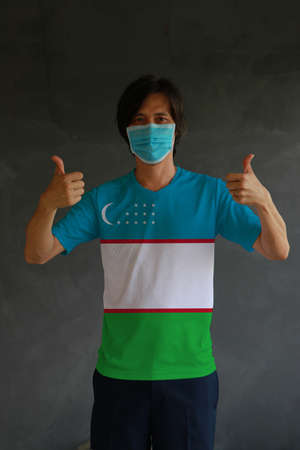 Man wearing hygienic mask and wearing Uzbek flag colored shirt with thumbs up on both hands. Concept of protect tiny dust or disease of Uzbekistan.の写真素材