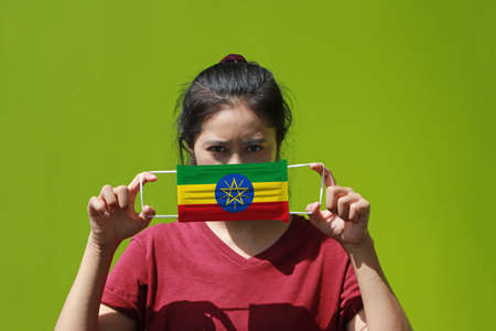 A woman and hygienic mask with Ethiopia flag pattern in her hand and raises it to cover her face on green background. A mask is a very good protection from Tiny Particle or respiratory disease.の写真素材