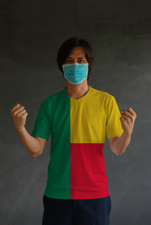 Man wearing hygienic mask and wearing Benin flag colored shirt and standing with raised both fist on dark wall background. Concept of protect tiny dust or disease.の写真素材