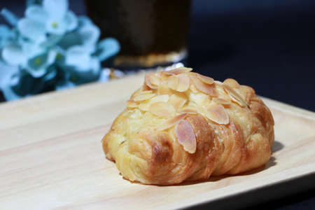 Almond flavor croissant placed on the square wooden tray with out focus tea in a glass and blue flowers on dark background. It is a French crescent shaped roll made of sweet flaky pastry, often eaten for breakfast.の写真素材