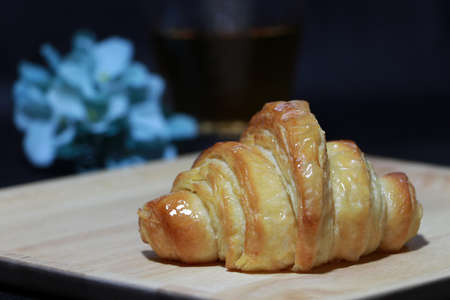 The croissant placed on the square wooden tray with out focus tea in a glass and blue flowers on dark background. It is a French crescent shaped roll made of sweet flaky pastry, often eaten for breakfast.の写真素材