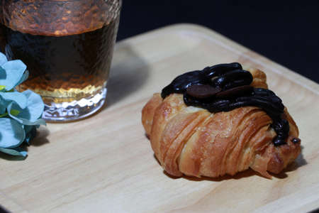 Chocolate flavor croissant placed on the square wooden tray with tea in a glass and blue flowers on dark background. It is a French crescent shaped roll made of sweet flaky pastry, often eaten for breakfast.の写真素材