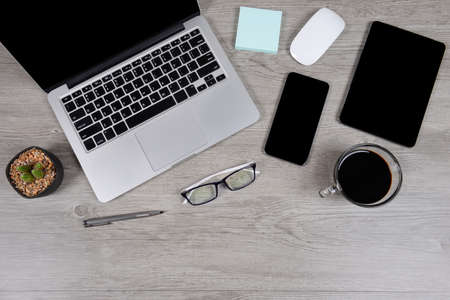 Office table with laptop computer, digital tablet, pen, smartphone, mouse, eyeglasses and coffee on white wood background. Desktop office mockup concept.の写真素材