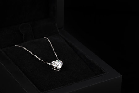 A heart shaped pendant with a chain is displayed in a black box. The pendant is made of clear glass and is set with diamonds. The box is black and the pendant is the main focus of the imageの写真素材