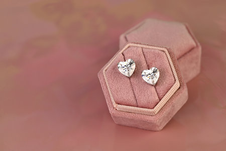 A pair of diamond earrings are displayed in a pink velvet box. The earrings are heart-shaped and are set in a clear, shiny material. The box is placed on a pink surface, which adds a touch of eleganceの写真素材