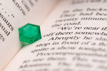 A green gemstone is on top of a page in a book. The gemstone is off to the side of the page, and the...