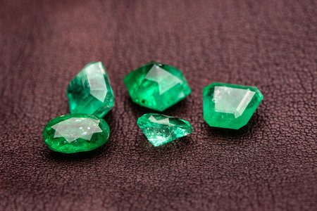A collection of green gemstones are displayed on a brown surface. The stones are of various shapes...