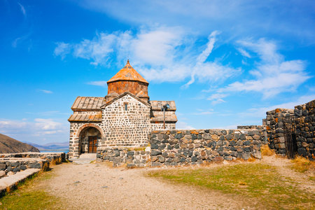 A small church Sevan Monastery Sevanavank with a steeple sits on a hillside. The sky is clear and...
