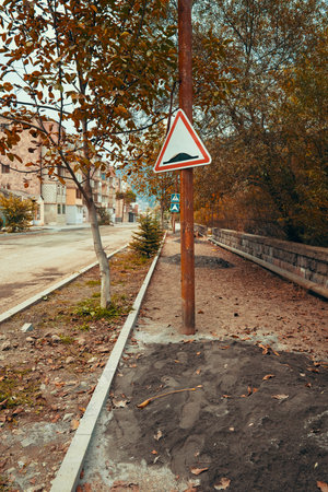 A sign on a pole in a city street warns of a steep drop. The street is lined with trees and has a sidewalkの写真素材
