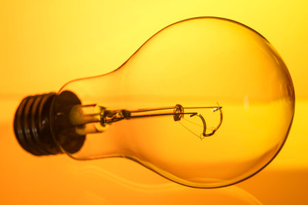 A light bulb is shown in a close up, with the bulb's filament visible. The bulb is yellow and is on...