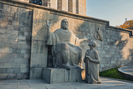 Yerevan Armenia 11.20.2025. The front of a large building matenadaran Mesrop Mashtots Institute of Ancient Manuscripts. A statue of a Mesrop Mashtots sitting on a pedestal with a woman kneeling in front of him. The statue is of a religious figure and the のeditorial素材