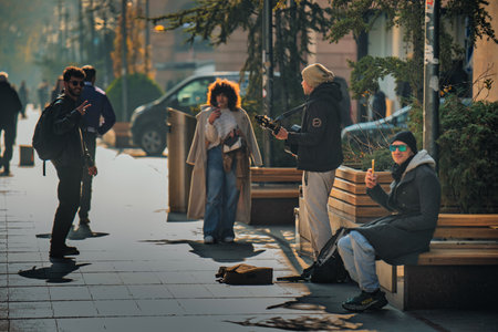 Yerevan Armenia 11.20.2025. A group of people are sitting on benches on a sidewalk. One man is playing guitar. A woman is holding a cameraのeditorial素材
