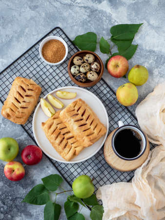 Top view food photography. Baked puff pastry crispy apple puff with ingredients and cup of coffee on a grey table. Puff pastry pies with apples, brown sugar, eggs and towel.の写真素材