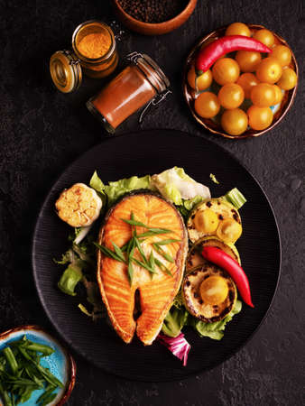 Healthy and diet food concept. Grilled salmon steak and vegetables on the grill on black background. Vertical top view, close-up. Red fish with garlic, yellow tomato and spicies on black plate.の写真素材