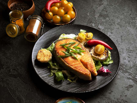 Red fish with garlic, yellow tomato and spicies on black plate. Healthy and diet food concept. Grilled salmon steak and vegetables on the grill on black background. Closeup. Soft focusの写真素材