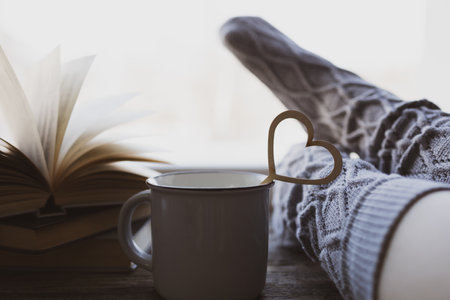 Woman resting keeping legs in warm socks with morning coffee and books. Lonely Valentine's Day. Love at distance, loneliness in self-isolation in the time of coronavirus. Selective focus on heart.の写真素材