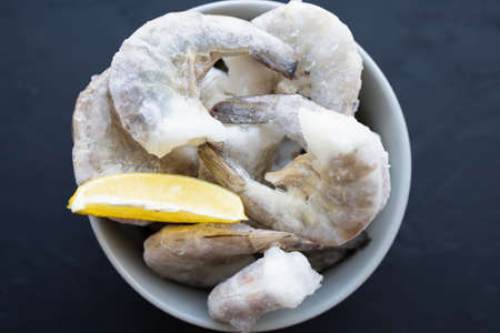Frozen big shrimps in grey bowl on black background with ice, lemon for cooking. Delicious king prawn without head for meal preparation. Enjoy natural seafood with low fat and high protein. Soft focusの写真素材