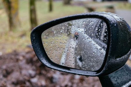 Close up of car side rear view mirror with rain drops on blurred background. Rainy day on the road....