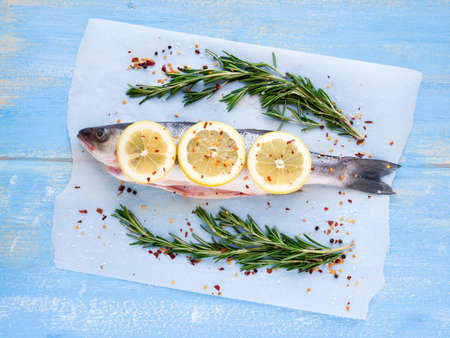 Mediterranean seafood concept. Raw seabass with lemon, spices and rosemary on the parchment paper on light blue wooden table. Fresh organic fish. Top view. Colorful and stylish composition.の写真素材