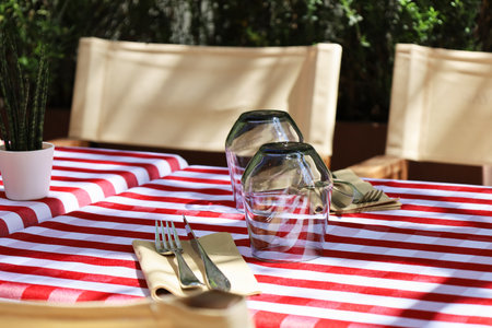 A served table with white and red tablecloth, two glasses and tableware is waiting for customers. Outdoor empty coffee terrace. Green cafe terrace on the pedestrian street in Europe.の写真素材