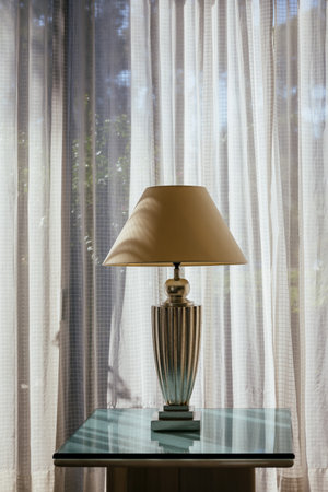Elegant white antique table lamp with fabric lampshade on the glass table near the window. Lamp silhouette on the curtain in sunny day. Classic decoration for home or hotel architectureの写真素材