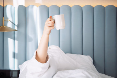A sleepy young woman with cup of coffee lying in bed with white cotton bedclothes. Concept of early morning wake up. Minimalism. Sunday loneliness relaxing, hygge mood.の写真素材