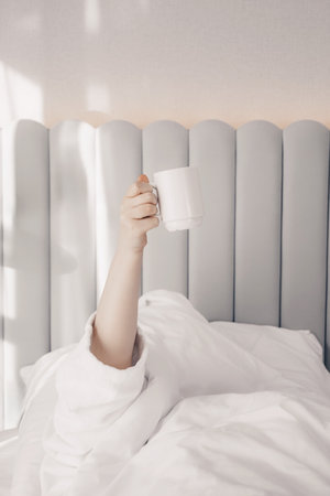 A sleepy young woman with cup of coffee lying in bed with white cotton bedclothes. Concept of early morning wake up. Pastel colors. Minimalism. Sunday loneliness relaxing, hygge mood.の写真素材