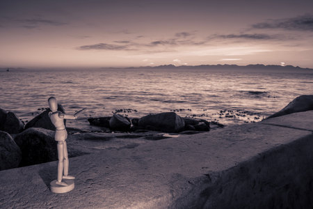 Wooden Human Manikin posing on beach at sunrise with a view of False Bay on a beautiful cloudy...