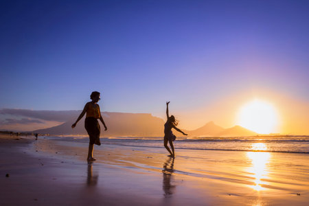 Two African women dancing on the beach at sunset, with Table Mountain and Cape Town in the...