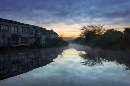 A row of houses surrounding a canal filled with reflections of the houses at sunrise on a misty morning, Cape Town, South Africaの写真素材
