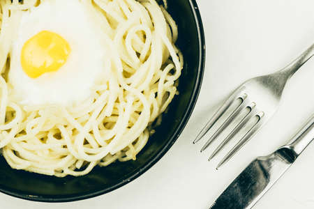 Fried egg and spaghetti in a black frying pan, fork and knife. Lunch in the cafe on a wooden table.の写真素材