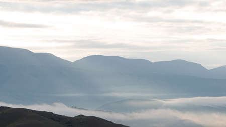 clouds and rays of dawn in mountain valley, soft light in early morning, meditation in nature. Gentle hills in bluish haze, silhouettes of mountainsの写真素材