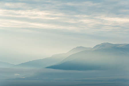clouds and rays of dawn in mountain valley, soft light in early morning, meditation in nature. Gentle hills in bluish haze, silhouettes of mountainsの写真素材