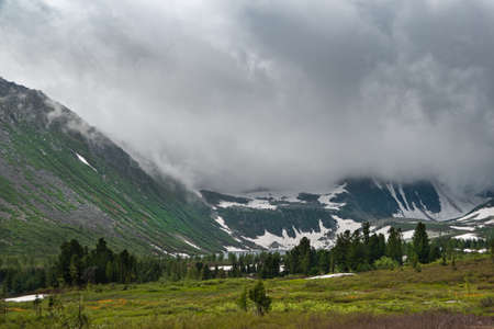Dense fog roofed tops of snow peaks, reducing visibility. Rain clouds over mountain valley. Slopes are covered with forest and glacier. Travel through spring highlands on foot.の写真素材