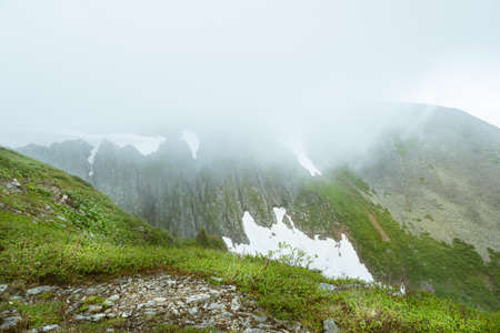 Dense fog roofed tops of snow peaks, reducing visibility. Rain clouds over mountain valley. Slopes are covered with forest and glacier. Travel through spring highlands on foot.の写真素材