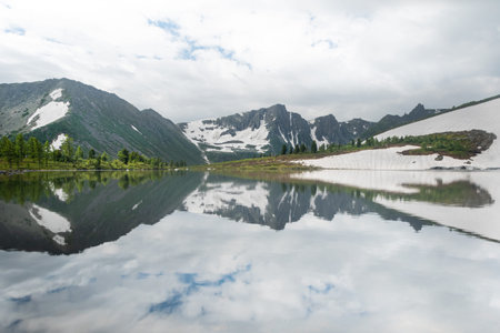 Mountain valley with snow, forest and lake at foot. Rocks are reflected in the river. Melting...