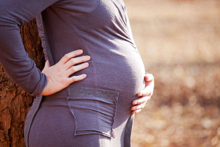 Young pregnant Caucasian woman leaning against a tree while holding her back and bellyの写真素材