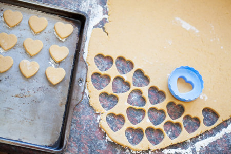 Blue plastic heart shaped cookie cutter and raw dough cookies on metal baking tray