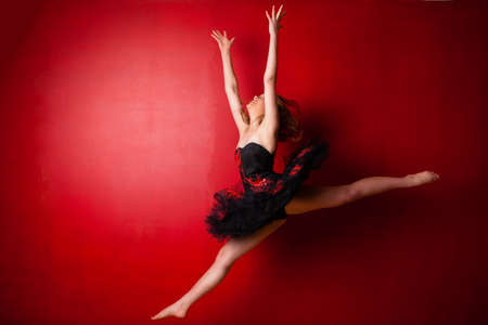Young Caucasian ballerina executing a jump against bright red wall, throwing her hands in the airの写真素材