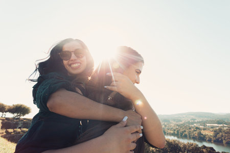 A couple of women having fun during a sunny day hugging each other while one smiles to camera with...