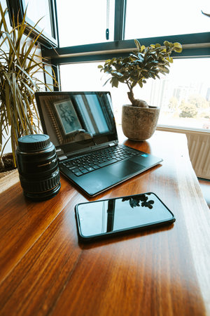 A work space composition with a black mobile phone, a plant, a photography lens and a black laptop