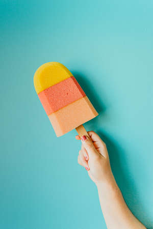 A super minimalistic hand grabbing a colorful foam ice cream in front of a pastel blue wallの写真素材