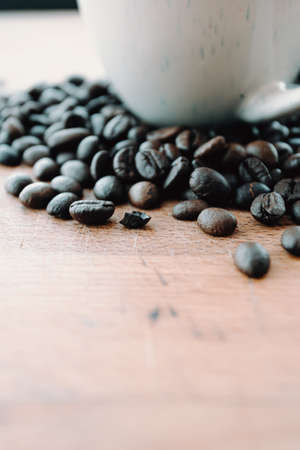 A super close up of some grains of coffee near a white cup and over a wood tableの写真素材