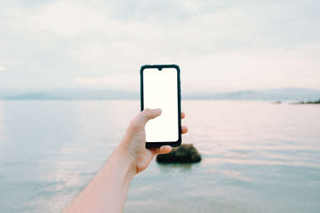 A young male hand grabbing a phone with copy space in vertical with the ocean as the backgroundの写真素材